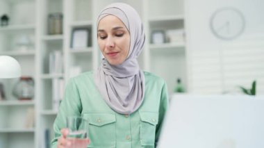 Ofisteki dizüstü bilgisayarda çalışırken camdan temiz su içen Müslüman iş kadınını kapatın. Gülümseyen girişimci işçiler sağlık dengesini kapalı tutuyor.