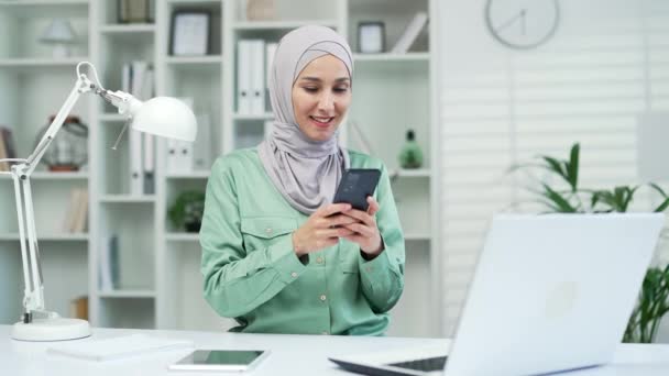 若いかなりイスラム教徒のアラビア語のビジネス女性は スマートフォンの入力を使用してチャットスクロールサーフィンウェブ職場で携帯電話を保持します屋内ヒジャーブの幸せな女性従業員の労働者は アプリのテキストメッセージを使用 — ストック動画