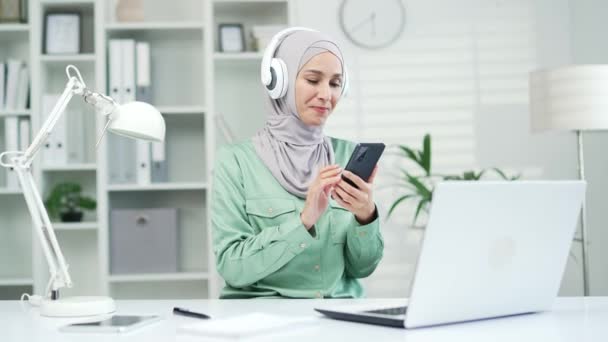 イスラム教徒の若い女性は 現代のオフィスでヘッドフォンで音楽やラジオを聞いてスマートフォンアプリの従業員を使用しています職場で楽しむヒジャーブの快適な女の子学生労働者屋内でプレイリスト電話で — ストック動画
