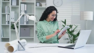 Genç kadın mimar yaratıcı tasarımcı çizimleri olan dijital tablet bloknotları kullanarak çalışıyor. Ofisteki İspanyol kadın apartman veya ev inşaatı ve ev yenileme tasarımları yapıyor.