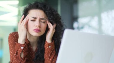 Kapatın. Ofiste otururken dizüstü bilgisayarla çalışırken baş ağrısı çeken esmer bir kadın. Üzgün kadın masaj yapıyor ve elleri kapalı şekilde şakaklarını ovuyor.