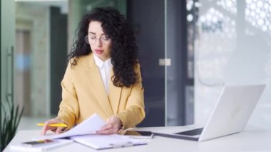 Gülümseyen esmer kadın finansör, modern ofiste işyerinde otururken iyi mali sonuçların incelemesinden memnun. Finansal raporlar tutan mutlu kadın kutlamaları