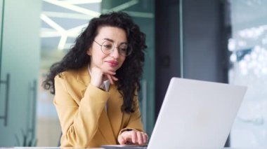 Gülümseyen kıvırcık esmer kadın, modern ofisteki çalışma masasında otururken bilgisayarını kullanarak flört sitesine giriyor. Takım elbiseli ve gözlüklü mutlu bir kadın bir mesaj yazar, internette sohbet eder.