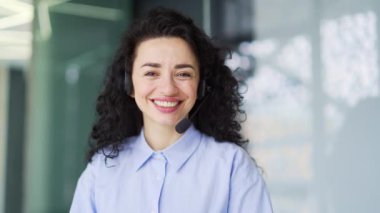 Kablosuz kulaklık takan esmer kadının portresi. Ofiste otururken görüntülü konuşma yapan müşteri hizmetleri operatörü. Çağrı merkezinin gülümseyen temsilcisi müşteriye kameraya bakmasını tavsiye eder.