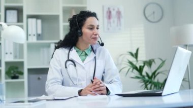 Kablosuz kulaklıklı kendine güvenen kadın doktor modern hastane kliniğinde dizüstü bilgisayarla konuşuyor. Tıp doktoru, ofisteki bir masada oturan bir hastaya danışır.