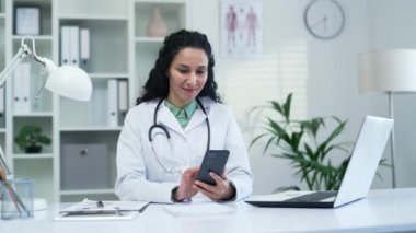 Beyaz önlüklü mutlu kadın doktor akıllı telefon kullanıyor, modern hastane kliniğinde mesaj yazıyor. Gülümseyen bir tıp doktoru bir hastaya ofisteki bir masada otururken mesaj atıyor.
