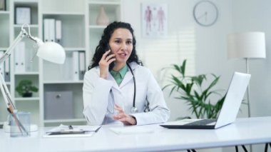 Beyaz önlüklü gülümseyen kadın doktor modern hastane kliniğinde akıllı telefondan konuşuyor. Esmer hekim bir konuşma yapar. Ofiste oturan bir hastaya danışır.
