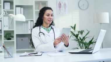 Beyaz önlüklü, gülümseyen kadın doktor modern hastane kliniğinde tablet kullanarak görüntülü konuşma yapıyor. Esmer bir hekim, ofisteki bir masada oturan bir hastaya danışıyor.