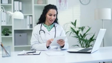 Beyaz önlüklü gülümseyen kadın doktor, modern bir hastane kliniğinde bir tablette daktilo kullanıyor. Tıp doktoru, iş yerinde otururken bir hastayla sohbet ediyor.