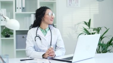 Mutlu gülümseyen beyaz önlüklü bayan doktor çalışma arasında koltukta dinleniyor modern hastane kliniğinde çalışma masasında oturuyor. Neşeli esmer tıp doktoru dinleniyor.