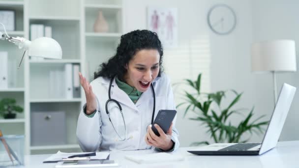 快乐兴奋的女医生坐在现代医院诊所的办公桌前 一边在智能手机上看到好消息 令人惊喜的黑发医生庆祝执政成功 — 图库视频影像
