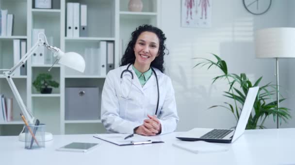 在现代医院诊所 身穿白衣的积极女医生一边微笑一边挥手看着相机 一边在办公桌前打招呼 友善的黑发女医务工作者医生 — 图库视频影像