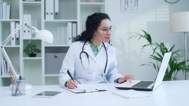 一名身穿白衣 面带微笑的女医生坐在一家现代医院诊所的办公桌前 一边看笔记本电脑 一边开处方 女医务工作者医生在办公室工作 — 图库视频影像