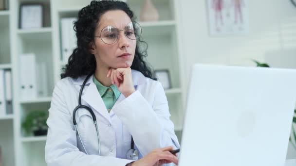靠近点一名严肃的女医生正在使用 她坐在医院诊所工作场所的办公桌前 在笔记本电脑上打字 一位黑发女性医务工作者的医生在办公室的电脑上工作 — 图库视频影像