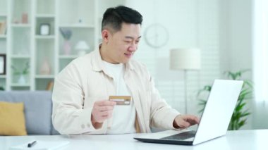Mutlu Asyalı adam internet üzerinden alışveriş yapıyor. Ev ofisinde otururken kredi kartı numarasını bilgisayara giriyor. Gülümseyen bir erkek satın alma ve ürün için promosyon fiyatından tatmin olur.