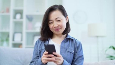 Evdeki oturma odasında kanepede otururken akıllı telefon kullanan Mutlu Asyalı kadını kapat. Gülümseyen kadın sosyal ağlara takılır, çevrimiçi mağazalardan alışveriş yapar, arkadaşlarıyla sohbet eder, mesajlar yazar.