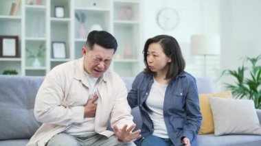 Hasta Asyalı adam evdeki kanepede otururken kalp krizi geçiriyor. Kadın kocasına destek oluyor ve telefonla ambulans çağırıyor. Erkek zor nefes alıyor, göğsünü tutuyor.