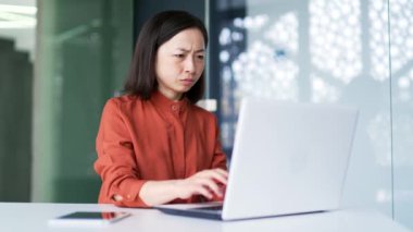 Hayal kırıklığına uğramış Asyalı kadın modern ofiste otururken dizüstü bilgisayar programının kötü performansından şikayet ediyor. İş kadını bilgisayarın, internetin ya da servisin çalışmasından memnun değil