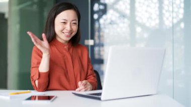 Modern ofiste iş yerinde otururken videoda konuşan mutlu Asyalı kadın. Gülümseyen dişi elini sallayarak selamlar, bir arkadaşıyla çevrimiçi bir konuşma yapar, uzaktan iletişim kurar.