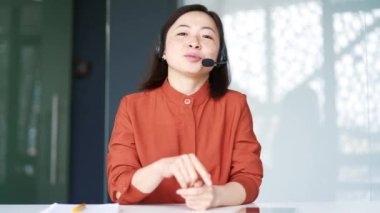 Webcam görüntüsü. Kulaklık takan Asyalı arkadaş canlısı bir kadın ofiste otururken videolu görüşme yapıyor. Çağrı merkezinin gülümseyen bir temsilcisi müşteriye tavsiyelerde bulunur ve soruları cevaplar.