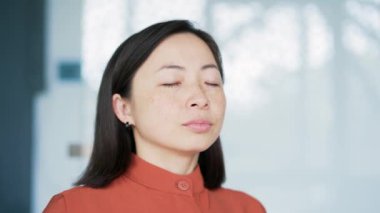 Modern bir ofiste gözleri kapalı dinlenen Asyalı bir kadın. Sakin, mutlu, dinlenen, derin nefes alan bir kadının portresi. Pozitif çalışan işe ara verdi. Meditasyon kavramı