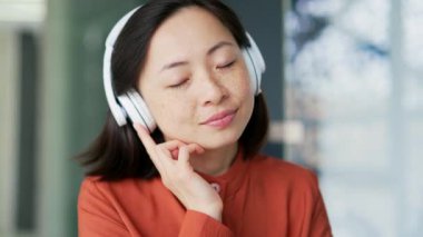 Kablosuz kulaklıklı tatmin olmuş Asyalı kadın modern ofiste otururken gözleri kapalı rahatlatıcı müzik dinliyor. Mutlu sakin kadın rahatlamanın sesine ve doğanın gürültüsüne alışır.
