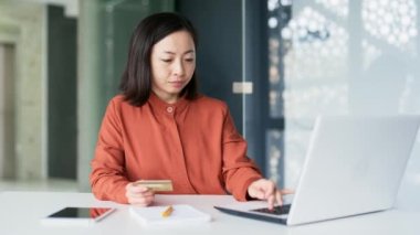Mutlu Asyalı iş kadını, modern ofisteki çalışma masasında otururken bilgisayarında kredi kartı numarasını yazarak online alışveriş yapıyor. Gülümseyen ve tatmin olmuş kadın çalışan satın alıyor.