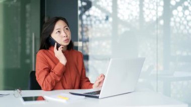 Müşteriyle akıllı telefonda konuşan ve iş yerinde masa başında otururken dizüstü bilgisayar kullanan meşgul bir iş kadını. Kendine güveni olan, tişörtlü, modern ofiste çalışan, bilgisayarda yazan kadın işçiler.