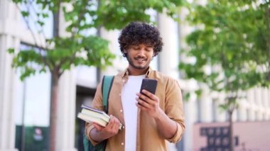 Sırt çantası ve kitapları olan yakışıklı gülümseyen öğrenci üniversite binasının yanındaki kampüs alanında akıllı telefon kullanıyor. Erkek sohbetleri, arkadaşlarıyla yazışmalar, sosyal ağlara göz atmalar.