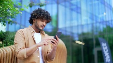 Bir ofis binasının yanındaki bankta otururken akıllı telefondan harika haberler okuyarak başarıyı kutlayan mutlu genç adam. Gülümseyen erkek neşeli bir mesaj aldı. Zaferin el hareketini gösteriyor.