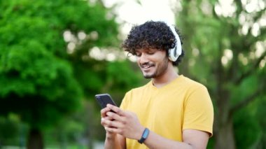 Kulaklıklı gülümseyen genç adam şehir parkındaki ağaçların arkasında müzik dinlemekten zevk alıyor. Mutlu erkek sporcu ya da sarı tişörtlü koşucu antrenmandan sonra dinleniyor.