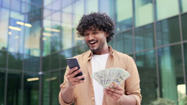 オフィスビルの近くに立っている間 若い幸せなビジネスマンはドル札のファンを振っている 手にスマートフォンや銀行券を持ち カメラを見ている興奮した男性を笑顔 金融の成功の概念 — ストック動画