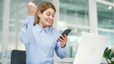 Mutlu iş kadını, modern ofisteki işyerinde otururken akıllı telefondan harika haberler okuyor. Gülümseyen ve memnun bir bayan çalışan keyifli bir mesaj bildirimi yapıyor.