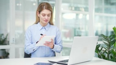 Bir kadın, iş yerinde oturan dizüstü bilgisayara bakarak not defterindeki video arama eğitim notlarını izliyor. Kadın çevrimiçi çalışıyor, uzaktan ders dinliyor, webinar, seminer, ders