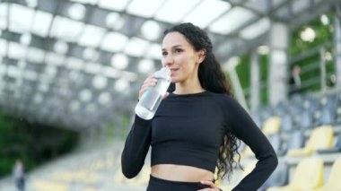 Şehir stadyumunda dikilirken şişeden su içen genç atletik kadın. Eşofmanlı esmer kadın spor yaptıktan sonra rahatlar, susuzluğunu ferahlatıcı bir içecekle giderir.