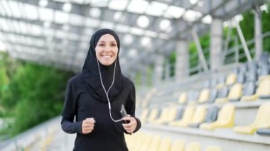 Şehir stadyumunda koşan, kulaklıklı, tesettürlü müslüman bir kadın. Mutlu sporcu kadın, akıllı telefon konsepti spor ve sağlıklı yaşam tarzı kullanarak dışarıda koşmaktan hoşlanıyor.