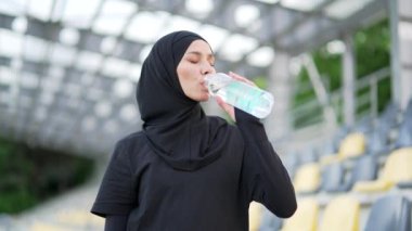 Şehirli bir stadyumda dikilirken şişeden su içen tesettürlü müslüman bir kadın. Mutlu, gülümseyen bir dişi antrenmandan sonra dinlenir, susuzluğunu ferahlatıcı temiz bir içecekle giderir.