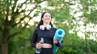 Elinde hasırla gülümseyen genç sporcu şehir parkındaki açık hava eğitimine gidiyor. Siyah eşofmanlı mutlu iyimser kadın yoga ya da jimnastik kursuna gidiyor.