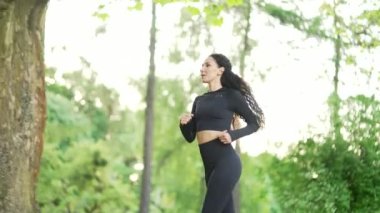 Yan görüş. Şehir parkında koşan gülümseyen genç bir spor kadını. Siyah eşofmanlı güzel bir esmer kadın sabah koşusunu doğayla iç içe yapıyor. Sağlıklı bir sporcu kadın antrenmanla meşgul.