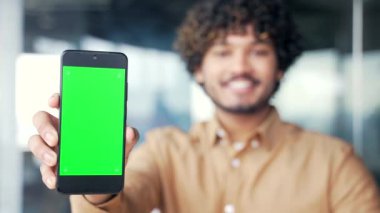 Modern ofiste oturan genç iş adamını gösteren yeşil ekranlı akıllı telefonu kapat. Renk anahtarı, reklam için dikey şablon düzeni. Gülümseyen adam cep telefonunu işaret ediyor.