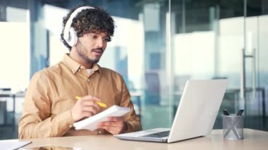 Kulaklıklı genç bir çalışan, ofiste dizüstü bilgisayara bakan bir not defterindeki video konuşma eğitim notlarını izliyor. Adam online çalışıyor, uzaktan bir kurs dinliyor, webinar, seminer