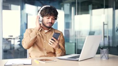 Kablosuz kulaklıklı genç, yakışıklı bir çalışan modern ofiste çalışırken müzik dinliyor. Mutlu bir işçi müzik listesindeki ritimle dans eder.