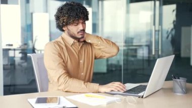 Modern ofisteki çalışma masasında otururken dizüstü bilgisayarla çalışırken boynu ağrıyan genç bir çalışan. Üzgün işçi masajları ve acı veren kaslar. Çok yorgun ve çok çalışıyor.