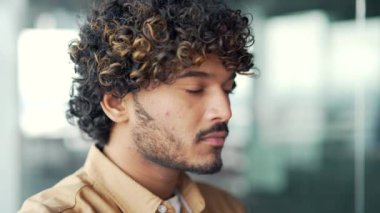 Modern ofisteki kendine güvenen yakışıklı genç adamın yakın portresi. Kameraya ciddi ciddi bakan kıvırcık saçlı, tişörtlü, düşünceli bir erkek çalışan. Yetişkin girişimcinin baş fotoğrafı.