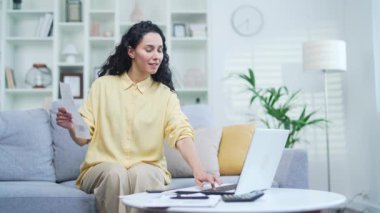 Evdeki koltukta otururken internet sitesi ya da uygulaması kullanarak ev finansmanı ile ilgilenen genç bir kadın, bir fatura tutuyor ve verileri bilgisayara girerek internet üzerinden ödeme formu dolduruyor.