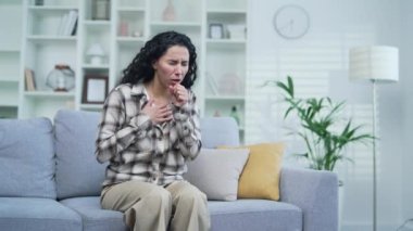 Evdeki oturma odasında kanepede oturan hasta genç bir kadın öksürüyor. Günlük giysiler içindeki yorgun kadın esmer elini göğsünde tutuyor. Soğuk algınlığı, virüs, bronşit veya grip belirtileri gösteriyor.