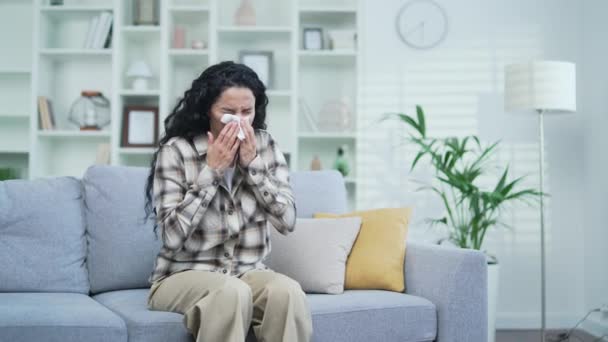 自宅のリビングルームでソファに座っている間 アレルギースニーズを持つ若い悲しいブルネットの女性 病気の女性はハンカチで鼻を拭く 彼女は風邪 ウイルス インフルエンザの最初の兆候を持っています — ストック動画