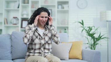 Genç esmer kadın evdeki oturma odasında kanepede otururken baş ağrısından muzdarip. Üzgün kadın elleriyle şakaklarına masaj yapıyor. Yorgun, acı çekiyor, migreni var ve tedaviye ihtiyacı var.