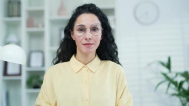 Evinde gözlüklü gülümseyen genç bir kadının portresini yakından çek. Kameraya bakarak poz veren dost canlısı bir kadın. Sarı gömlekli mutlu öğretmen oturma odasında.