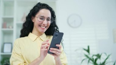 Kapatın. Evdeki oturma odasında akıllı telefon kullanan mutlu, genç esmer kadın. Gözlüklü gülümseyen bir kadın sosyal ağlara takılır, çevrimiçi mağazalardan alışveriş yapar, arkadaşlarıyla sohbet eder, mesajlar yazar.